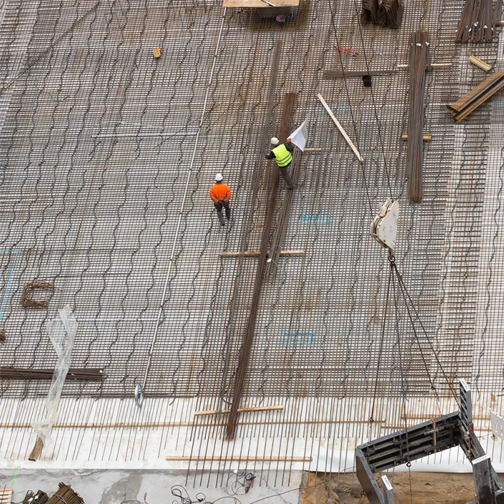 Overhead view of a construction site with two workers in safety helmets walking on a steel reinforcement grid with materials and equipment nearby.
